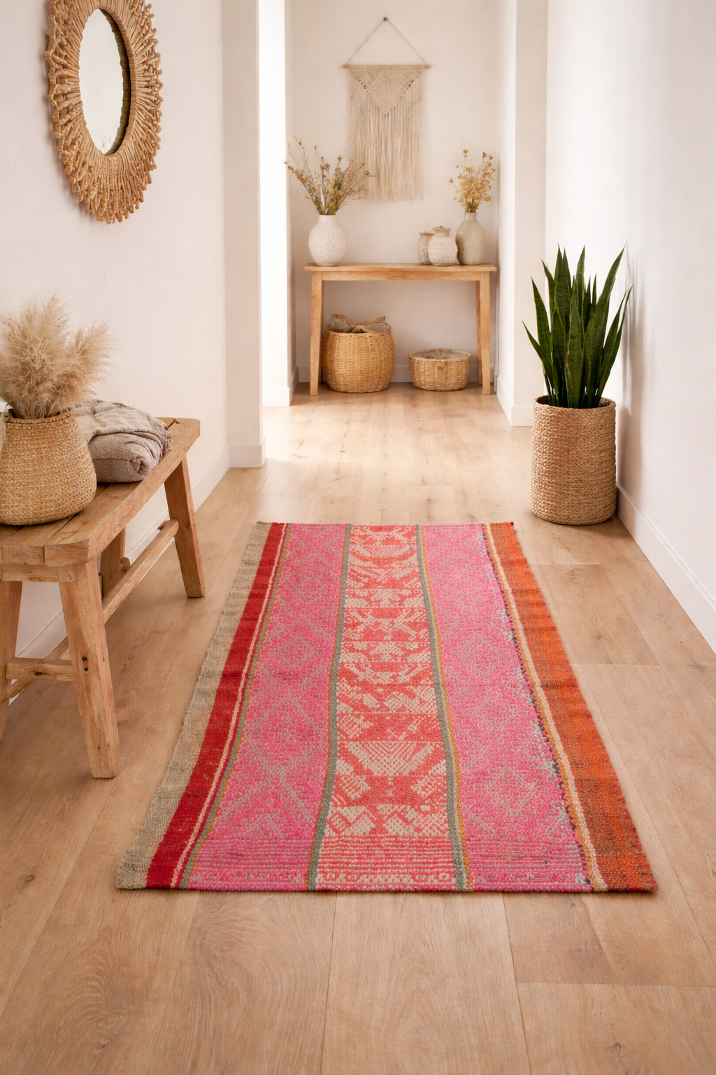 Heller Boho-Flur mit hellem Holzboden. Ein peruanischer Frazada-Teppich mit asymmetrischem Muster in Pink, Rot und Orange liegt mittig. Holzbank, Pflanze, Keramik und Makramee ergänzen den Raum. Natürliches Licht schafft eine warme Atmosphäre.